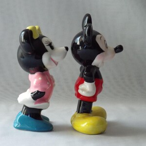 Puede incluir: Dos figuras de cer&aacute;mica de Mickey y Minnie Mouse. Mickey lleva pantalones cortos rojos, zapatos amarillos y guantes blancos. Minnie lleva un vestido rosa con lunares blancos, zapatos azules y un lazo amarillo.