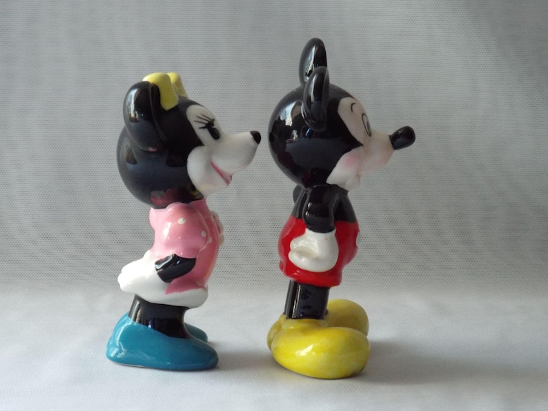 Puede incluir: Dos figuras de cer&aacute;mica de Mickey y Minnie Mouse. Mickey lleva pantalones cortos rojos, zapatos amarillos y guantes blancos. Minnie lleva un vestido rosa con lunares blancos, zapatos azules y un lazo amarillo.