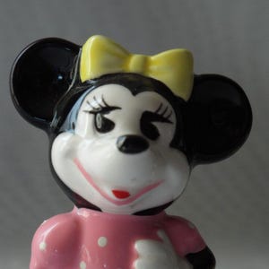 Puede incluir: Figura de cer&aacute;mica de Minnie Mouse. La figura tiene orejas negras, un lazo amarillo y un vestido rosa con lunares blancos. La cara es blanca con ojos negros, pesta&ntilde;as y boca roja.