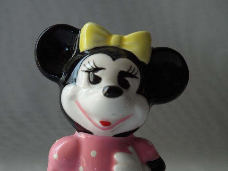 Puede incluir: Figura de cer&aacute;mica de Minnie Mouse. La figura tiene orejas negras, un lazo amarillo y un vestido rosa con lunares blancos. La cara es blanca con ojos negros, pesta&ntilde;as y boca roja.