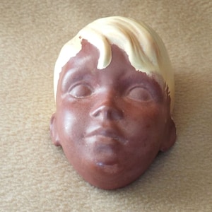 May include: A ceramic sculpture of a child's head with a brown face and blonde hair. The sculpture is painted in shades of brown and yellow.