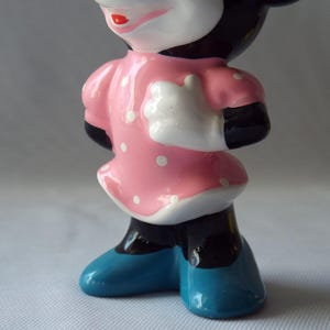 Puede incluir: Figura de cer&aacute;mica de un personaje de dibujos animados, con un vestido rosa con lunares blancos, brazos y piernas negros, guantes blancos y zapatos azules. El personaje tiene una cara blanca, labios rojos y orejas negras.