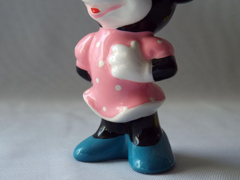 Puede incluir: Figura de cer&aacute;mica de un personaje de dibujos animados, con un vestido rosa con lunares blancos, brazos y piernas negros, guantes blancos y zapatos azules. El personaje tiene una cara blanca, labios rojos y orejas negras.
