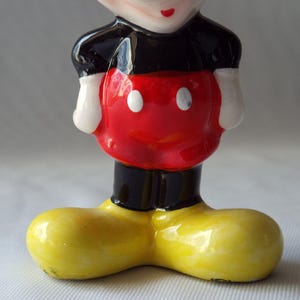 Puede incluir: Figura de cer&aacute;mica de Mickey Mouse. La figura tiene un torso rojo con dos botones blancos, pantalones cortos negros, guantes blancos y zapatos amarillos grandes. La cara es visible, con una cabeza negra, boca roja y ojos blancos.