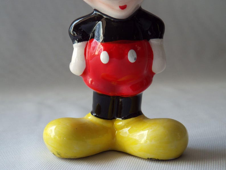 Puede incluir: Figura de cer&aacute;mica de Mickey Mouse. La figura tiene un torso rojo con dos botones blancos, pantalones cortos negros, guantes blancos y zapatos amarillos grandes. La cara es visible, con una cabeza negra, boca roja y ojos blancos.