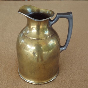 May include: A small, polished brass jug with a dark grey handle. The jug has a rounded body and a flared lip.