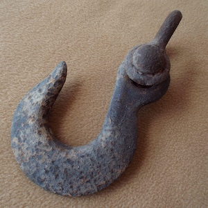 May include: A weathered, dark gray metal hook with a curved design. The hook has a textured surface and a rounded top with a bolt. The item appears to be vintage or antique.