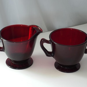 May include: A ruby red glass creamer and sugar bowl set. The creamer features a single handle and a pouring spout, while the sugar bowl has two handles. Both pieces are made of translucent red glass and sit on a circular pedestal base.