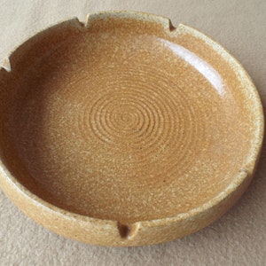 May include: A round, ceramic ashtray in a speckled tan colour. The ashtray has a wide rim with three notches and a single cigarette rest. The interior features concentric circular grooves.
