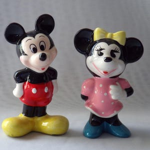 Puede incluir: Dos figuras de cer&aacute;mica de Mickey Mouse y Minnie Mouse. Mickey lleva una camisa negra, pantalones cortos rojos y zapatos amarillos. Minnie viste un vestido rosa con lunares blancos, un lazo amarillo y zapatos azules.