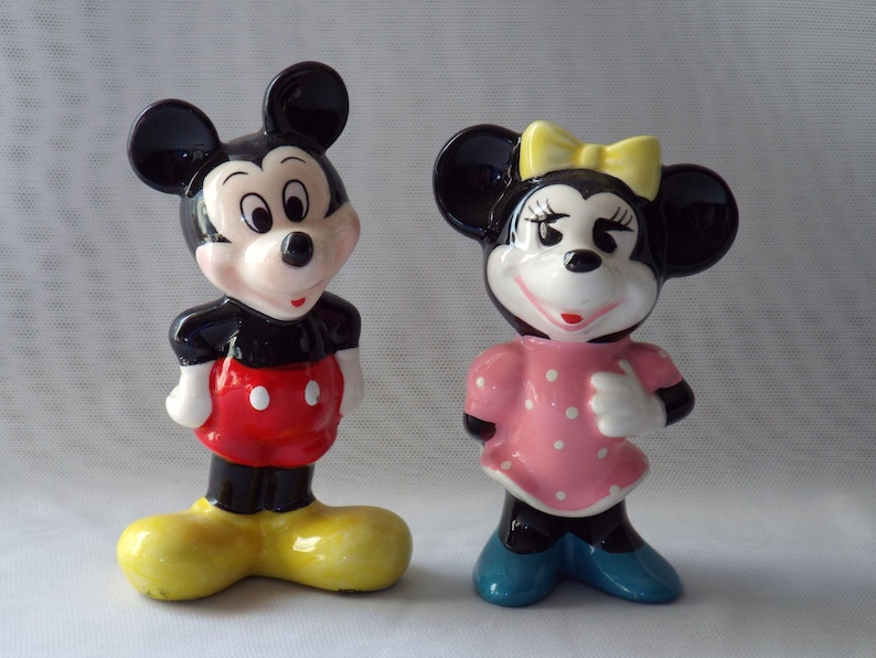 Puede incluir: Dos figuras de cer&aacute;mica de Mickey Mouse y Minnie Mouse. Mickey lleva una camisa negra, pantalones cortos rojos y zapatos amarillos. Minnie viste un vestido rosa con lunares blancos, un lazo amarillo y zapatos azules.