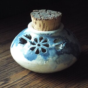 May include: A small, white ceramic jar with a blue glaze and a cork lid. The jar has a decorative cutout design of flowers.