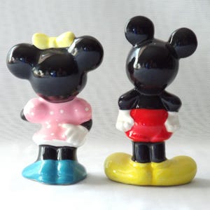 Puede incluir: Dos figuras de cer&aacute;mica de personajes de dibujos animados. Una es una figura negra con un lazo amarillo, un vestido rosa con lunares blancos y zapatos azules. La otra es una figura negra con pantalones cortos rojos, guantes blancos y zapatos amarillos.