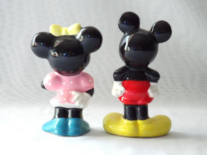 Puede incluir: Dos figuras de cer&aacute;mica de personajes de dibujos animados. Una es una figura negra con un lazo amarillo, un vestido rosa con lunares blancos y zapatos azules. La otra es una figura negra con pantalones cortos rojos, guantes blancos y zapatos amarillos.