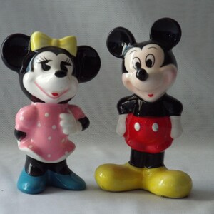 Puede incluir: Dos figuras de cer&aacute;mica de Mickey y Minnie Mouse. Minnie lleva un vestido rosa con lunares blancos, un lazo amarillo y zapatos azules. Mickey lleva pantalones cortos rojos con botones blancos y zapatos amarillos.