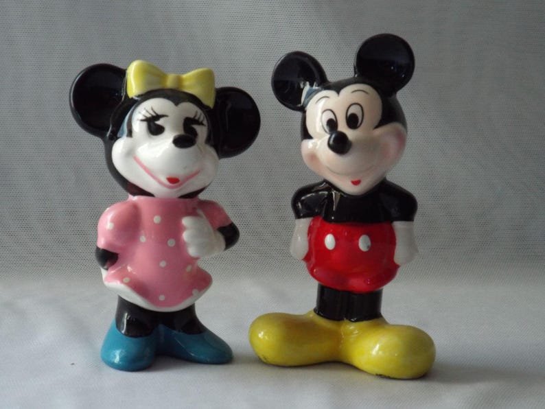 Puede incluir: Dos figuras de cer&aacute;mica de Mickey y Minnie Mouse. Minnie lleva un vestido rosa con lunares blancos, un lazo amarillo y zapatos azules. Mickey lleva pantalones cortos rojos con botones blancos y zapatos amarillos.
