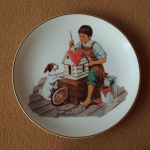 May include: A white ceramic plate with a gold rim features a painting of a boy and a dog painting a small house. The boy is wearing a red and blue shirt and green pants. The dog is white with brown spots. The painting is signed "Norman Rockwell".