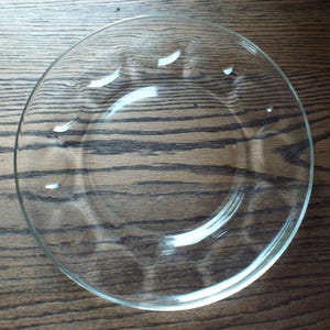 May include: A clear glass plate with a simple design. The plate is round with a slightly raised edge and small decorative indentations. The plate is sitting on a dark wooden surface.