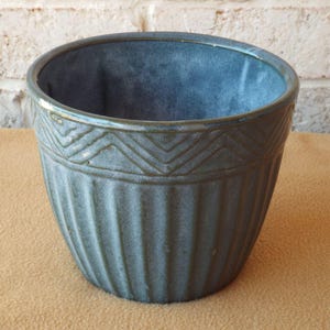 May include: A blue-green ceramic planter with a textured, ribbed exterior and a decorative geometric band near the rim. The planter has a round shape and is suitable for indoor or outdoor use. The background features a brick wall.