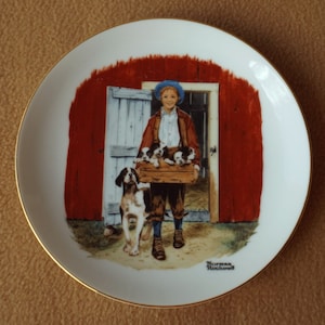 May include: A white ceramic plate with a gold rim features a painting of a young boy holding a basket of puppies. The boy is wearing a blue hat, a red and white shirt, and blue jeans. The background is a red barn door with a white door. The painting is signed "Norman Rockwell".