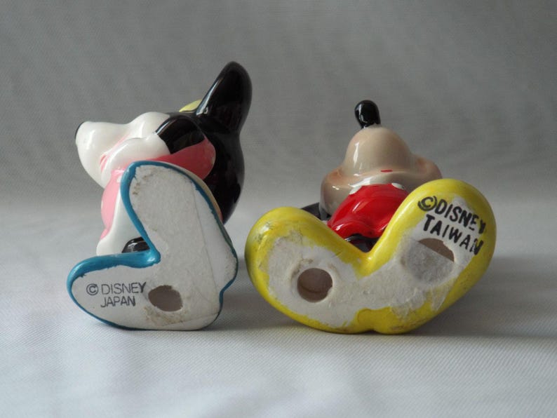 Puede incluir: Dos figuras vintage de Disney. Una es negra, blanca y rosa, con una base azul y el texto "Disney Japan". La otra es amarilla, roja y marr&oacute;n claro, con el texto "Disney Taiwan".