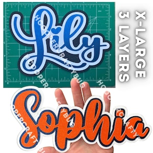 May include: Two layered name cutouts. The top cutout displays the name Lily in blue shades on a green cutting mat with a 6-inch width. The bottom cutout displays the name Sophia in orange and dark blue glitter, labeled as X-LARGE 3 LAYERS.