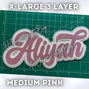 May include: A three-layer custom name cutout featuring the name Aliyah in glittery medium pink script, layered over white and silver glitter cardstock. The text X-LARGE 3 LAYER and MEDIUM PINK is visible, along with a green cutting mat grid.