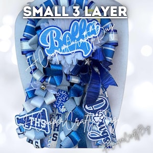 May include: A small 3-layer homecoming mum featuring blue and silver ribbons, a white flower base, and glittery blue cutouts with the text Bella, THS, and Dawgs. The decorative accessory includes layered loops, streamers, and a small silver star.