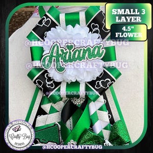 May include: A homecoming mum featuring a 4.5-inch white flower, green and black ribbons with heart patterns, and a green glitter cutout reading Ariana. The design includes layered ribbons, a megaphone accent, and text stating SMALL 3 LAYER.