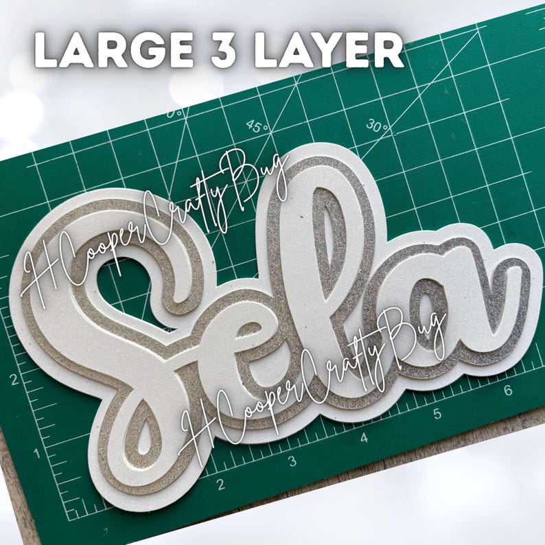 May include: A large 3-layer cutout of the name Sela in a cursive script font. The design features alternating layers of white and silver glitter material, creating a dimensional effect. The text LARGE 3 LAYER is visible at the top of the image.