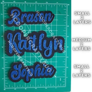 May include: Three custom name cutouts featuring blue glitter script lettering layered over black backing. The names Brason, Kaitlyn, and Sophia are displayed on a green cutting mat with inch markings, labeled as small or medium with 2 layers.