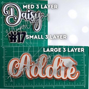May include: Three-layer custom name and number cutouts displayed on a green cutting mat with inch markings. The top section shows 'Daisy' and '#17' in navy and white glitter. The bottom section shows 'Addie' in white, orange, and silver glitter.
