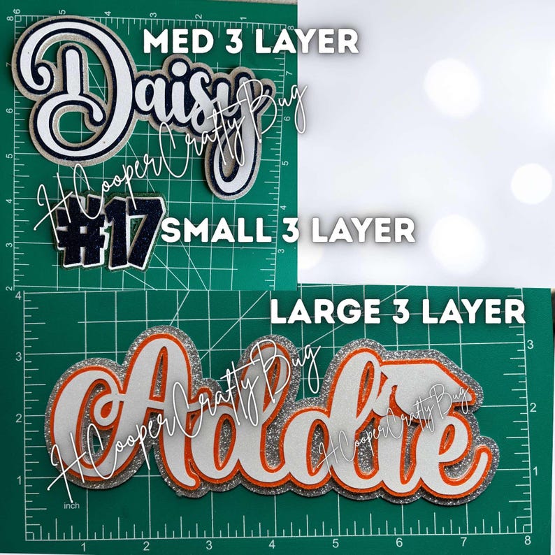May include: Three-layer custom name and number cutouts displayed on a green cutting mat with inch markings. The top section shows 'Daisy' and '#17' in navy and white glitter. The bottom section shows 'Addie' in white, orange, and silver glitter.