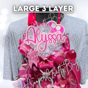 May include: A large 3-layer decorative badge featuring the name Alyssa in bright pink script. The design includes multiple layers of pink and glittery ribbons, a soccer ball graphic, and intricate embellishments arranged in a vertical display.