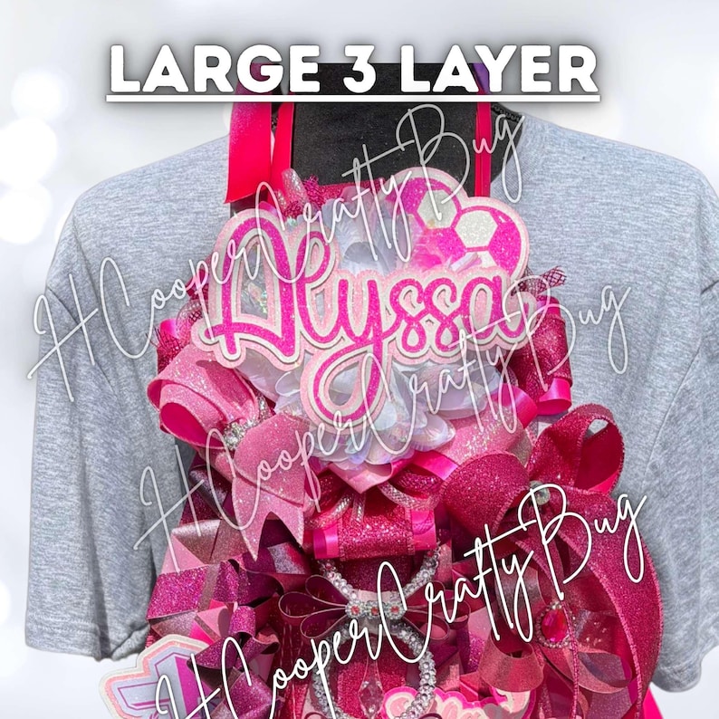 May include: A large 3-layer decorative badge featuring the name Alyssa in bright pink script. The design includes multiple layers of pink and glittery ribbons, a soccer ball graphic, and intricate embellishments arranged in a vertical display.