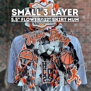 May include: A small 3-layer homecoming mum with a 5.5-inch flower and 12-inch skirt, featuring orange and black ribbons, basketball-themed cutouts, a basketball hoop, and the names Tyler and Riley in glittery orange script on a grey shirt.
