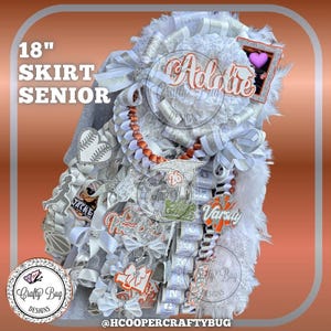May include: A senior skirt, approximately 46 cm long, in white, silver, and orange. The skirt features ribbons, bows, and embellishments, with the name "Addie" and the word "Varsity" visible. The design includes a heart and a baseball.