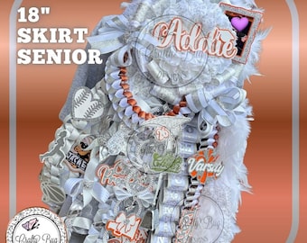 May include: A senior skirt, approximately 18 inches long, in white, silver, and orange. The skirt features ribbons, bows, and embellishments, with the name "Addie" and the word "Varsity" visible. The design includes a heart and a baseball.