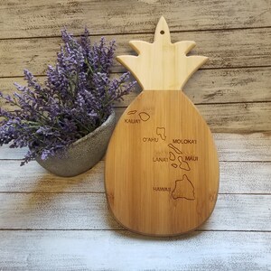 May include: A wooden pineapple-shaped cutting board with a map of the Hawaiian islands engraved on it. The islands are labeled with their names: Kauai, Oahu, Molokai, Lanai, Maui, and Hawaii.