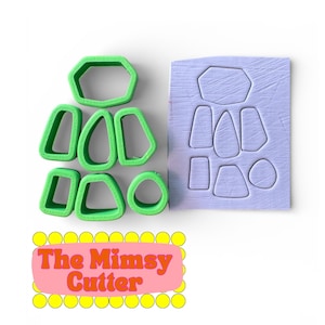 Op de afbeelding: Een set groene plastic koekjessnijders in verschillende geometrische vormen, waaronder een zeshoek, een cirkel en verschillende onregelmatige vormen. Er is ook een lichtpaarse mal met bijpassende vormen. De tekst "The Mimsy Cutter" staat op een roze banner.