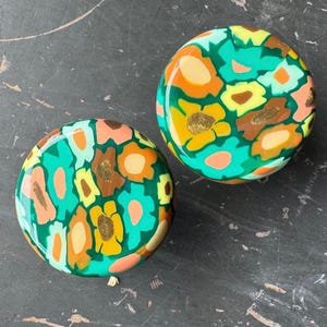 May include: Two circular decorative items with a floral design. The pattern incorporates green, orange, yellow, brown, and pink hues. The objects have a glossy finish and are set against a dark background.