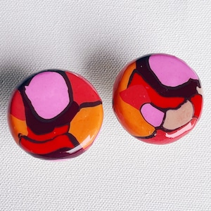 May include: A pair of round, colorful earrings with a swirling abstract pattern in shades of pink, red, orange, and brown.