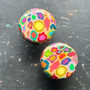 May include: Two round, cream-colored ornaments with a colorful, abstract floral pattern. The ornaments are painted with bright colors, including pink, yellow, green, blue, and orange.