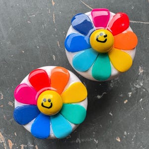 May include: Two colorful ceramic flower-shaped ornaments with a smiley face in the center. Each flower has six petals in different colors, including red, orange, yellow, green, blue, and purple.