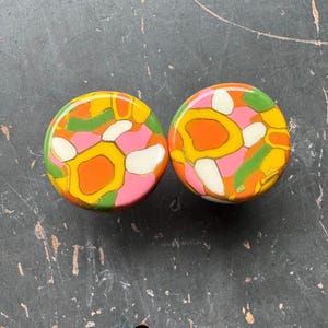 Peut inclure: Deux boutons ronds en céramique colorés avec un motif floral abstrait rose, orange, jaune, vert et blanc.