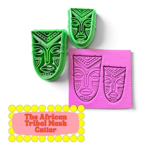 Op de afbeelding: Twee groene plastic koekjesvormpjes in de vorm van maskers met geometrische patronen, naast een roze vierkante plaat met twee afgedrukte maskerontwerpen. De tekst The African Tribal Mask Cutter staat in een roze kader.