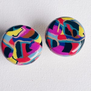 Puede incluir: Un par de pendientes redondos coloridos con un patrón geométrico en tonos de rosa, azul, amarillo, negro y marrón.