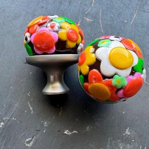 Może przedstawiać: Dwa ozdobne gałki do szafek z brązową podstawą i kolorowymi wzorami kwiatów. Gałki są wykonane z ceramiki i mają błyszczące wykończenie.