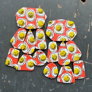 May include: Two multi-tiered dangle earrings featuring a vibrant orange background decorated with a repeating pattern of white and yellow fried egg motifs. Each egg includes a yellow centre with a black smiley face. The earrings consist of several geometric shapes connected by small gold-toned metal jump rings.