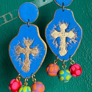 Könnte beinhalten: Blaue Ohrringe mit goldenem Kreuzdesign und bunten, baumelnden Perlen. Die Ohrringe haben eine blaue Oberseite und einen goldenen Rand. Die baumelnden Perlen sind pink, orange und blau mit Tupfen.